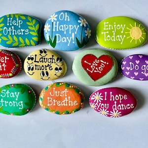 May include: A collection of ten smooth, hand-painted stones featuring vibrant colors and inspirational text. Messages include Help Others, Oh Happy Day, Enjoy Today, Never Quit, Laugh more, I Love You, Do good, Stay Strong, Just Breathe, and I hope you dance.