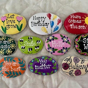May include: A collection of hand-painted decorative stones with inspirational messages and floral designs. Messages include "Let love grow," "Happy Birthday," "Here comes the sun," "You are magic," "Smile," "Hello," "Stay Wild," "You are the best," "You matter," and "Believe."