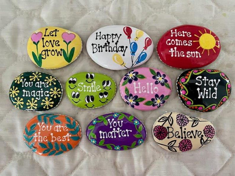May include: A collection of hand-painted decorative stones with inspirational messages and floral designs. Messages include "Let love grow," "Happy Birthday," "Here comes the sun," "You are magic," "Smile," "Hello," "Stay Wild," "You are the best," "You matter," and "Believe."
