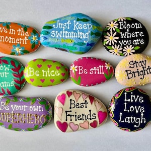 May include: A collection of ten smooth, hand-painted river stones featuring various colors and inspirational phrases including Live in the moment, Just keep swimming, Bloom where you are, Follow your dreams, Be nice, Be still, Stay Bright, Be your own SUPERHERO, Best Friends, and Live Love Laugh.