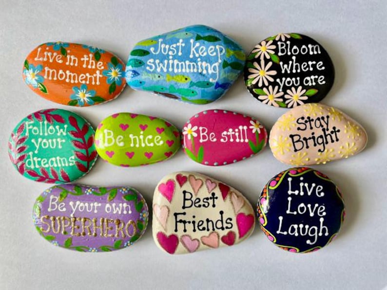 May include: A collection of ten smooth, hand-painted river stones featuring various colors and inspirational phrases including Live in the moment, Just keep swimming, Bloom where you are, Follow your dreams, Be nice, Be still, Stay Bright, Be your own SUPERHERO, Best Friends, and Live Love Laugh.