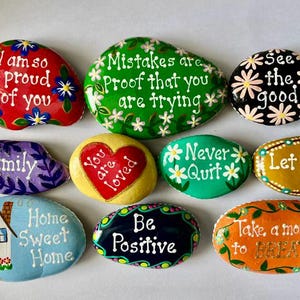 May include: A collection of ten smooth, hand-painted stones featuring various colors and inspirational messages including I am so proud of you, Mistakes are proof that you are trying, See the good, Family, You are loved, Never Quit, Let it go, Home Sweet Home, Be Positive, and Take a moment to BREATHE.