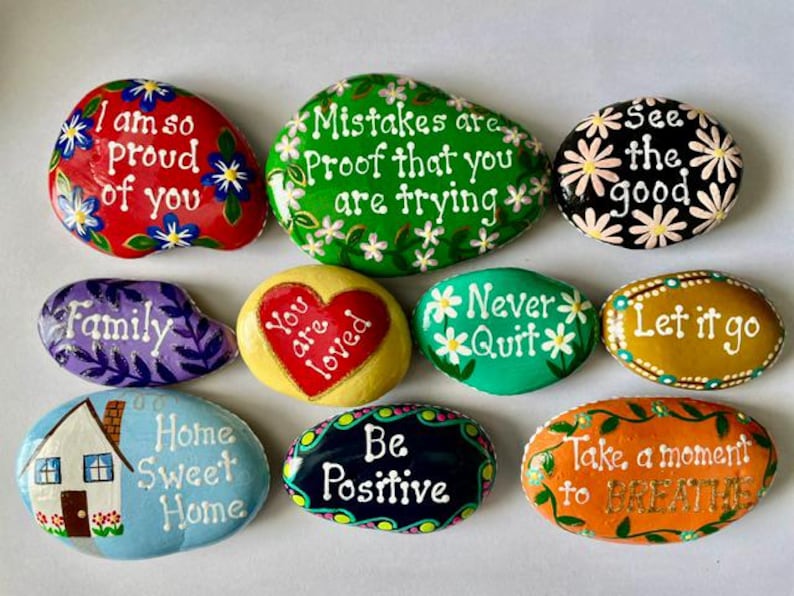 May include: A collection of ten smooth, hand-painted stones featuring various colors and inspirational messages including I am so proud of you, Mistakes are proof that you are trying, See the good, Family, You are loved, Never Quit, Let it go, Home Sweet Home, Be Positive, and Take a moment to BREATHE.