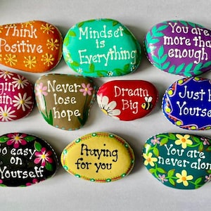 May include: A collection of ten smooth, hand-painted river stones featuring colorful backgrounds and white or black text. Phrases include Think Positive, Mindset is Everything, You are more than enough, Begin again, Never lose hope, Dream Big, Just be Yourself, Go easy on Yourself, Praying for you, and You are never alone.