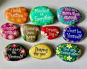 May include: A collection of ten smooth, hand-painted river stones featuring colorful backgrounds and white or black text. Phrases include Think Positive, Mindset is Everything, You are more than enough, Begin again, Never lose hope, Dream Big, Just be Yourself, Go easy on Yourself, Praying for you, and You are never alone.