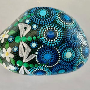 May include: A hand-painted, smooth river rock with a dark background. The rock features a design of blue and turquoise dots arranged in circular patterns. White daisy-like flowers and green leaves are painted on one side, adding a natural contrast.
