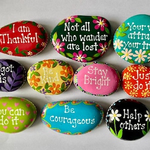 May include: A collection of nine smooth, hand-painted stones featuring colorful designs and inspirational text including I am Thankful, Not all who wander are lost, Your vibe attracts your tribe, You got this, Be Kind, Stay Bright, Just do it, You can do it, Be courageous, and Help others.