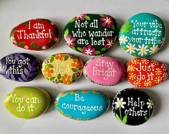 May include: A collection of nine smooth, hand-painted stones featuring colorful designs and inspirational text including I am Thankful, Not all who wander are lost, Your vibe attracts your tribe, You got this, Be Kind, Stay Bright, Just do it, You can do it, Be courageous, and Help others.