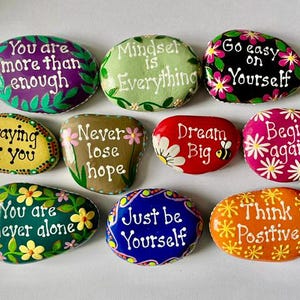 May include: A collection of nine smooth, hand-painted river stones featuring colorful backgrounds and white lettering. The stones display inspirational phrases including You are more than enough, Mindset is Everything, Go easy on Yourself, Praying for you, Never lose hope, Dream Big, Begin again, You are never alone, Just be Yourself, and Think Positive.