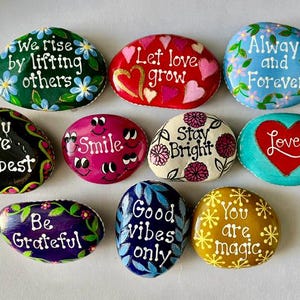 Pode incluir: Uma cole&ccedil;&atilde;o de dez pedras pintadas com mensagens inspiradoras. Cada pedra tem uma cor diferente e apresenta texto pintado &agrave; m&atilde;o e elementos decorativos como flores e cora&ccedil;&otilde;es. Mensagens: "We rise by lifting others" e "Love".