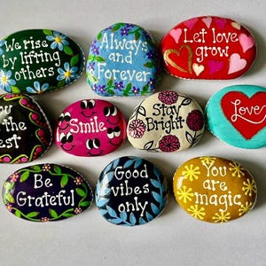 May include: A collection of ten smooth, hand-painted river stones featuring various colors and inspirational phrases including We rise by lifting others, Always and Forever, Let love grow, You are the best, Smile, Stay Bright, Love, Be Grateful, Good vibes only, and You are magic.
