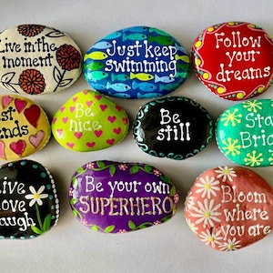 May include: A collection of hand-painted decorative stones with inspirational quotes. The stones are oval-shaped and feature phrases like "Live in the moment," "Best Friends," and "Bloom where you are." Each stone is painted with bright colors and floral or heart designs.