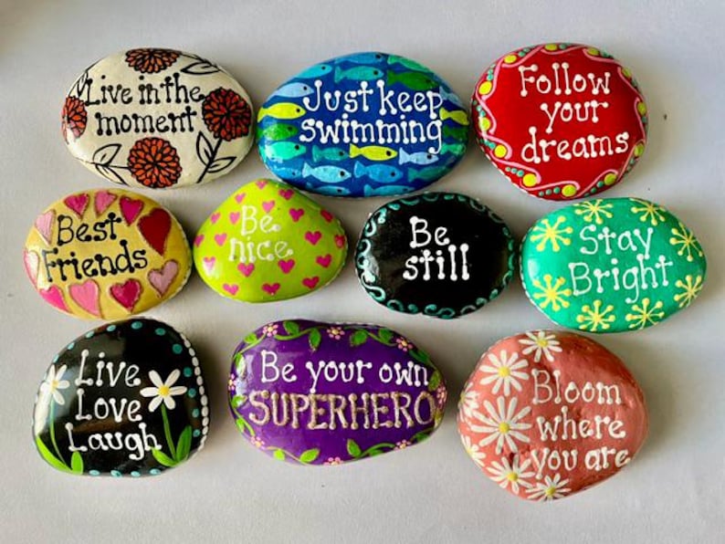 May include: A collection of hand-painted decorative stones with inspirational quotes. The stones are oval-shaped and feature phrases like "Live in the moment," "Best Friends," and "Bloom where you are." Each stone is painted with bright colors and floral or heart designs.