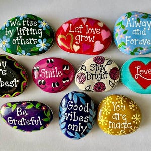 Pode incluir: Uma cole&ccedil;&atilde;o de pedras decorativas pintadas &agrave; m&atilde;o com mensagens inspiradoras. Cada pedra apresenta designs &uacute;nicos, incluindo padr&otilde;es florais, cora&ccedil;&otilde;es e frases inspiradoras como "Smile" e "Good Vibes Only."