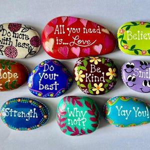 May include: A collection of ten smooth, hand-painted river stones featuring colorful designs and inspirational text including Do more with less, All you need is Love, Believe, Hope, Do your best, Be Kind, Smile, Strength, Why not?, and Yay You!