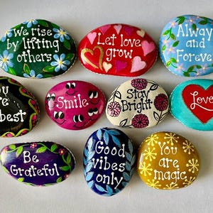 Pode incluir: Uma cole&ccedil;&atilde;o de pedras decorativas pintadas &agrave; m&atilde;o com mensagens inspiradoras. Cada pedra apresenta designs &uacute;nicos, incluindo padr&otilde;es florais, cora&ccedil;&otilde;es e afirma&ccedil;&otilde;es positivas como "Smile", "Love" e "Good Vibes Only". As pedras s&atilde;o em v&aacute;rias cores, incluindo verde, vermelho, azul e amarelo.