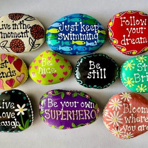 May include: A collection of hand-painted decorative rocks with inspirational quotes and floral designs. The rocks feature phrases like "Live in the moment," "Best Friends," and "Bloom where you are." Each rock is a unique piece of art with vibrant colors and detailed patterns.