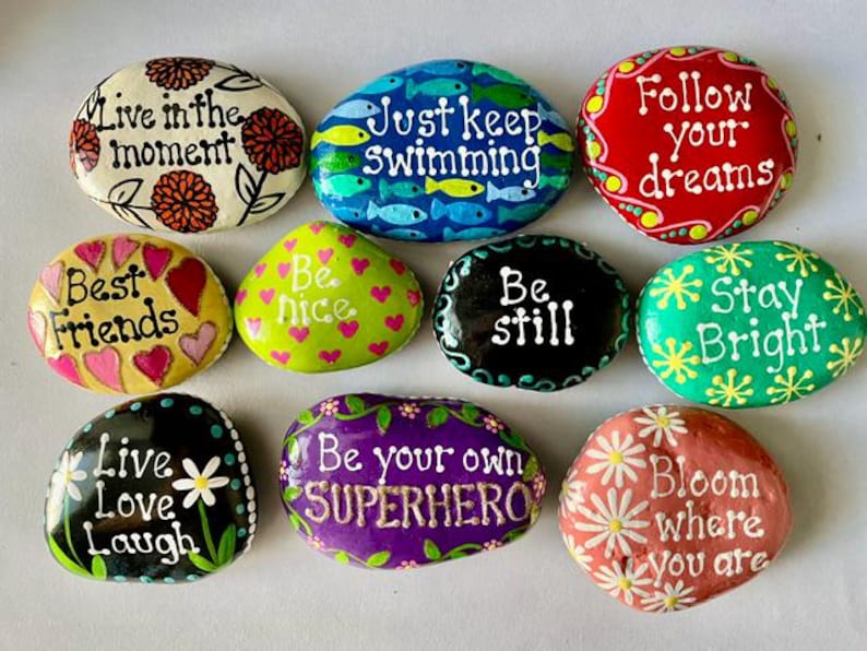 May include: A collection of hand-painted decorative rocks with inspirational quotes and floral designs. The rocks feature phrases like "Live in the moment," "Best Friends," and "Bloom where you are." Each rock is a unique piece of art with vibrant colors and detailed patterns.
