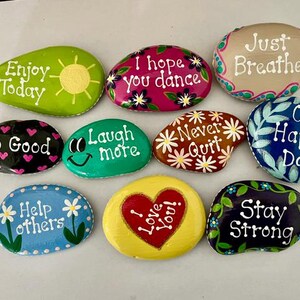 May include: A collection of hand-painted rocks with inspirational messages. Each rock is a different color and features phrases like "Enjoy Today," "I Love You," and "Stay Strong." The rocks are decorated with floral and heart designs.