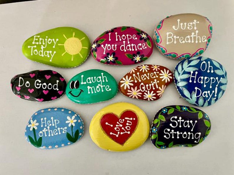 May include: A collection of hand-painted rocks with inspirational messages. Each rock is a different color and features phrases like "Enjoy Today," "I Love You," and "Stay Strong." The rocks are decorated with floral and heart designs.
