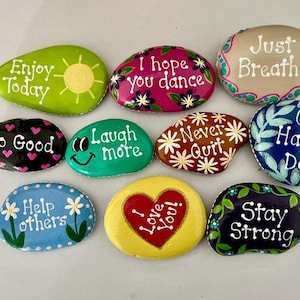 May include: A collection of painted rocks with inspirational messages. The rocks are various colors, including green, pink, blue, and yellow. Each rock features a different phrase, such as "Enjoy Today" and "Stay Strong", in white lettering.
