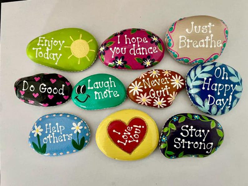 May include: A collection of painted rocks with inspirational messages. The rocks are various colors, including green, pink, blue, and yellow. Each rock features a different phrase, such as "Enjoy Today" and "Stay Strong", in white lettering.
