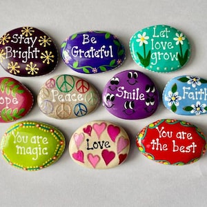 May include: A collection of nine smooth, oval-shaped river stones painted in various colors. Each stone features hand-painted text and designs, including phrases like Stay Bright, Be Grateful, Let love grow, Hope, Peace, Smile, Faith, You are magic, Love, and You are the best.