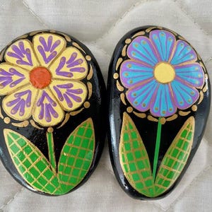 Pode incluir: Duas pedras pintadas com flores. Uma pedra tem uma flor amarela com detalhes roxos e folhas verdes. A outra pedra tem uma flor azul com detalhes roxos e folhas verdes. Ambas as pedras t&ecirc;m um fundo preto.
