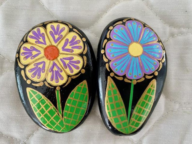 Pode incluir: Duas pedras pintadas com flores. Uma pedra tem uma flor amarela com detalhes roxos e folhas verdes. A outra pedra tem uma flor azul com detalhes roxos e folhas verdes. Ambas as pedras t&ecirc;m um fundo preto.