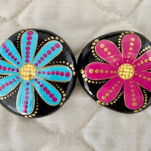 Pode incluir: Duas pedras pintadas com fundos pretos e flores coloridas. Uma pedra tem uma flor azul com pontos rosa e um centro dourado. A outra pedra tem uma flor rosa com pontos dourados e um centro dourado.