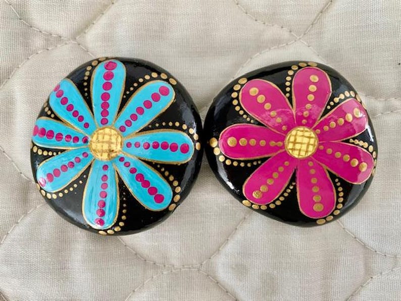 Pode incluir: Duas pedras pintadas com fundos pretos e flores coloridas. Uma pedra tem uma flor azul com pontos rosa e um centro dourado. A outra pedra tem uma flor rosa com pontos dourados e um centro dourado.