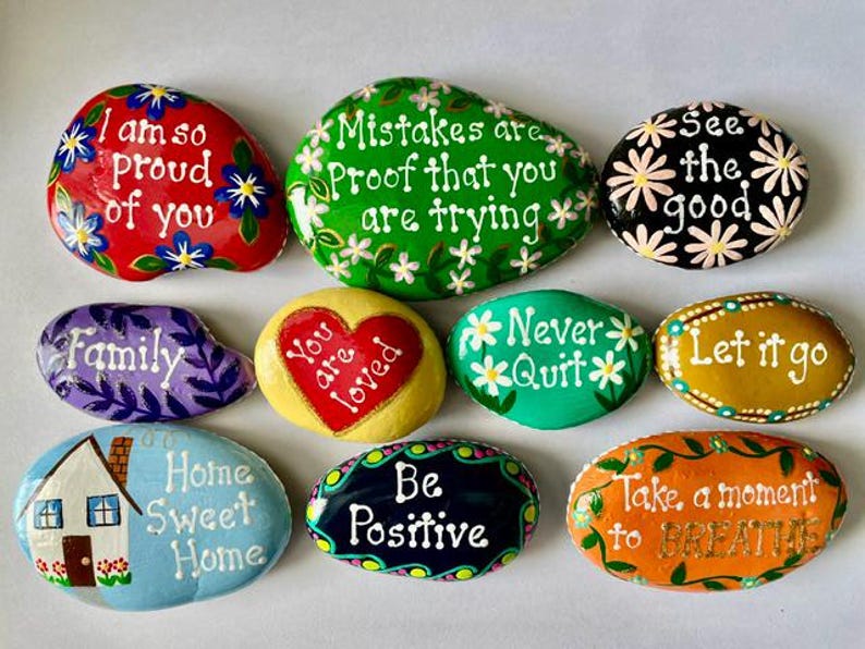 May include: A collection of ten smooth, hand-painted river stones featuring various colors and inspirational messages. Text includes "I am so proud of you," "Mistakes are proof that you are trying," "See the good," "Family," "You are loved," "Never Quit," "Let it go," "Home Sweet Home," "Be Positive," and "Take a moment to BREATHE."