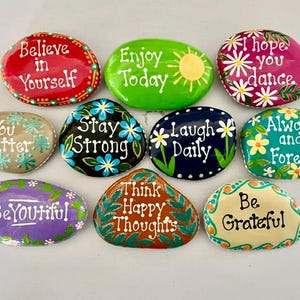 May include: A collection of ten smooth, hand-painted stones featuring colorful backgrounds and white text. Phrases include Believe in Yourself, Enjoy Today, I hope you dance, You matter, Stay Strong, Laugh Daily, Always and Forever, BeYOUTiful, Think Happy Thoughts, and Be Grateful.