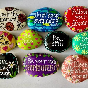 May include: A collection of hand-painted rocks with inspirational quotes and floral designs. The rocks feature phrases like "Live in the moment," "Just keep swimming," and "Follow your dreams." Colors include red, green, blue, and yellow.