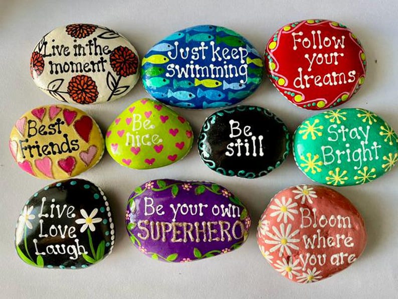 May include: A collection of hand-painted rocks with inspirational quotes and floral designs. The rocks feature phrases like "Live in the moment," "Just keep swimming," and "Follow your dreams." Colors include red, green, blue, and yellow.