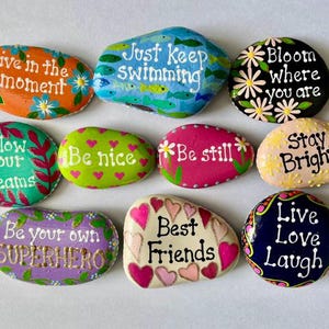 May include: A collection of ten smooth, hand-painted river stones featuring various colors and inspirational phrases including Live in the moment, Just Keep Swimming, Bloom where you are, Follow your dreams, Be nice, Be still, Stay Bright, Be your own SUPERHERO, Best Friends, and Live Love Laugh.