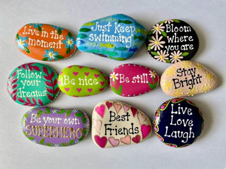May include: A collection of ten smooth, hand-painted river stones featuring various colors and inspirational phrases including Live in the moment, Just Keep Swimming, Bloom where you are, Follow your dreams, Be nice, Be still, Stay Bright, Be your own SUPERHERO, Best Friends, and Live Love Laugh.