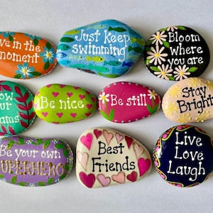 May include: A collection of ten smooth, hand-painted river stones featuring various colors and inspirational phrases. The stones display messages including Live in the moment, Just keep swimming, Bloom where you are, Follow your dreams, Be nice, Be still, Stay Bright, Be your own SUPERHERO, Best Friends, and Live Love Laugh.