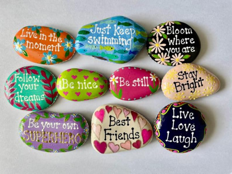 May include: A collection of ten smooth, hand-painted river stones featuring various colors and inspirational phrases. The stones display messages including Live in the moment, Just keep swimming, Bloom where you are, Follow your dreams, Be nice, Be still, Stay Bright, Be your own SUPERHERO, Best Friends, and Live Love Laugh.