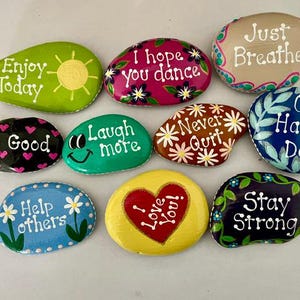 May include: A collection of painted rocks with inspirational messages. The rocks are various shapes and colors, including green, pink, and blue. Messages include "Enjoy Today," "I hope you dance," and "Stay Strong."