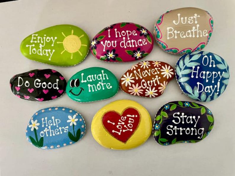 May include: A collection of painted rocks with inspirational messages. The rocks are various shapes and colors, including green, pink, and blue. Messages include "Enjoy Today," "I hope you dance," and "Stay Strong."