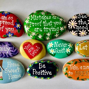 May include: A collection of nine smooth, hand-painted river stones featuring various colors and inspirational messages. The stones display phrases like I am so proud of you, Mistakes are proof that you are trying, See the good, Family, You are loved, Never quit, Let it go, Home Sweet Home, Be Positive, and Take a moment to BREATHE.