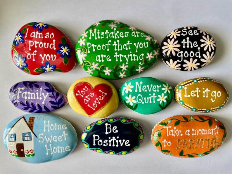 May include: A collection of nine smooth, hand-painted river stones featuring various colors and inspirational messages. The stones display phrases like I am so proud of you, Mistakes are proof that you are trying, See the good, Family, You are loved, Never quit, Let it go, Home Sweet Home, Be Positive, and Take a moment to BREATHE.
