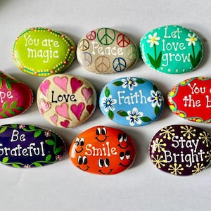 May include: A collection of nine smooth, hand-painted stones featuring colorful designs and inspirational text. Phrases include You are magic, Peace, Let love grow, Hope, Love, Faith, You are the best, Be Grateful, Smile, and Stay Bright.