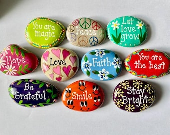 May include: A collection of nine smooth, hand-painted stones featuring colorful designs and inspirational text. Phrases include You are magic, Peace, Let love grow, Hope, Love, Faith, You are the best, Be Grateful, Smile, and Stay Bright.