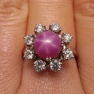 May include: A silver-toned ring featuring a central round cabochon-cut pink star sapphire gemstone surrounded by a halo of eight small, clear, round-cut diamonds arranged in a floral pattern on a polished metal band.