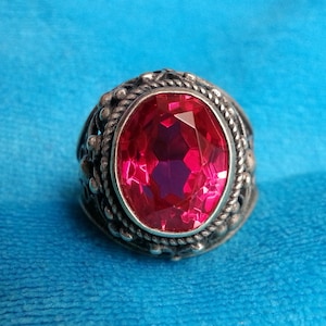 May include: A silver ring featuring a large, oval-cut, vibrant pink gemstone set in a decorative metal frame. The band and setting are crafted with intricate, textured metalwork, including a twisted rope border surrounding the central stone.