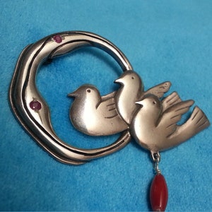 May include: Silver-toned brooch featuring three stylized doves within a crescent-shaped frame. The frame has three small red accents. A red bead hangs from the bottom. The brooch is set against a blue background.