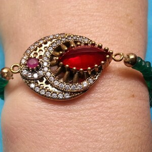 May include: A bracelet featuring a central gold-tone metal medallion with a marquise-cut red stone, a small round pink stone, and clear pavé crystals. The bracelet band consists of small green beads connected to the central decorative element.