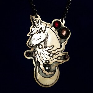 May include: A silver-toned metal pendant on a dark chain featuring an engraved unicorn head with a horn, flowing mane with star cutouts, and a crescent moon at the base. The design is accented with four small, round, dark red faceted gemstones.